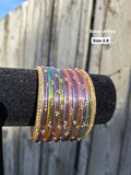 Stack of translucent multi-color pastel glass bangles (lavender, yellow, peach, green) with gold sequin accents and gold stone border bangles displayed on a black velvet bar outdoors.