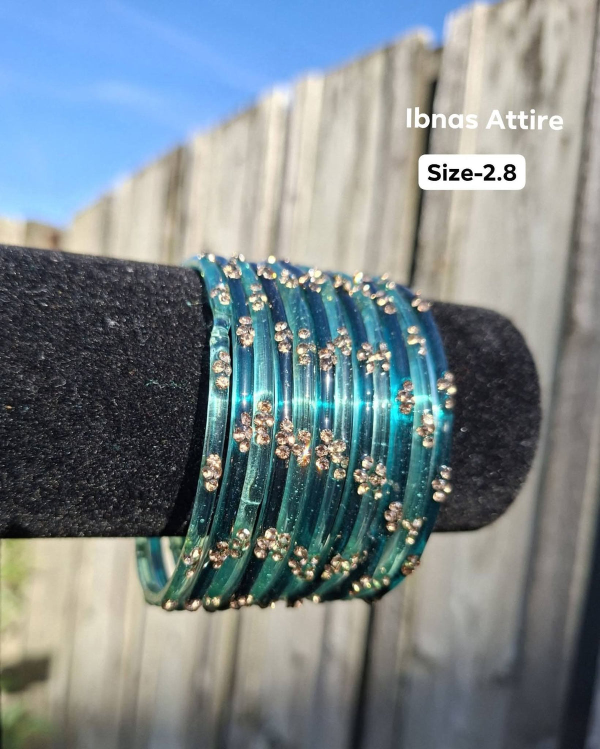 Stack of translucent aqua teal / ocean blue-green glass bangles with gold sequin accents displayed on a black velvet bar outdoors against a wooden fence.  Smooth glass construction that produces a traditional, melodious chime when worn.  