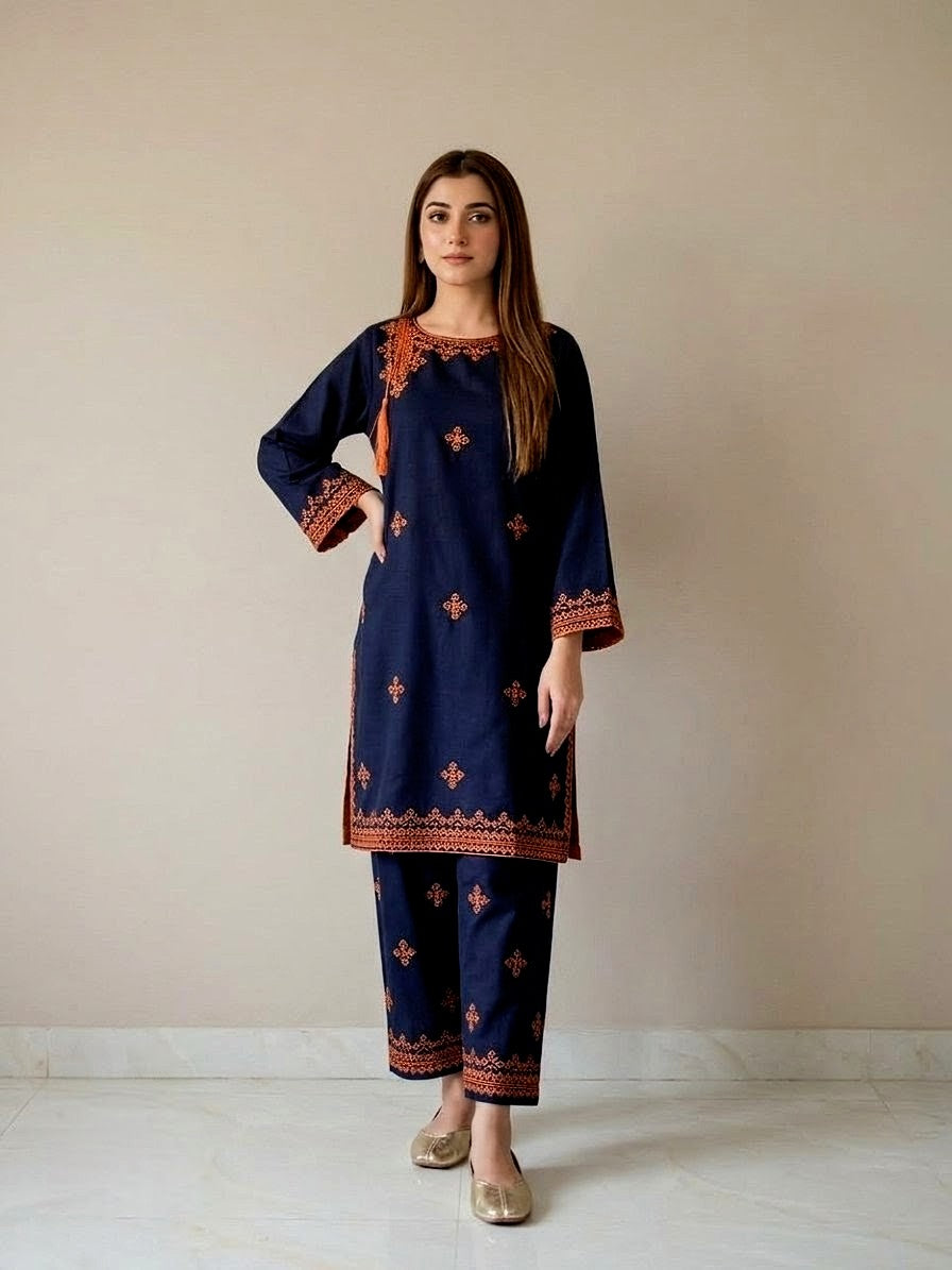 A woman modeling a navy blue pure pakistani summer lawn kurta and pant set with rust-orange border and floral embroidery, and mirror Detailing standing against a plain beige wall.