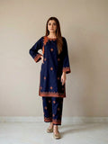 A woman modeling a navy blue pure pakistani summer lawn kurta and pant set with rust-orange border and floral embroidery, and mirror Detailing standing against a plain beige wall.
