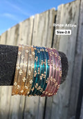 Stack of translucent glass bangles in champagne gold, peacock teal, and mauve pink with gold sequin accents displayed on a black velvet bar outdoors against a wooden fence.