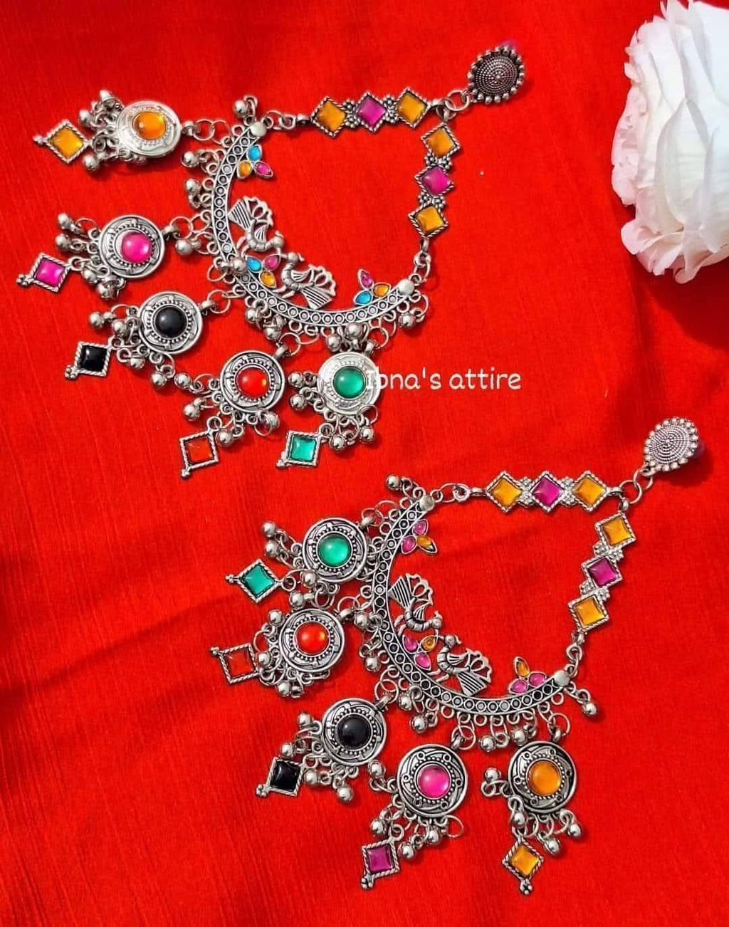 A pair of ornate silver-toned Indian chandbali earrings decorated with colorful rhinestones in pink, orange, yellow, green, black, and red, displayed on a bright red fabric background.