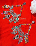 A pair of ornate silver-toned Indian chandbali earrings decorated with colorful rhinestones in pink, orange, yellow, green, black, and red, displayed on a bright red fabric background.