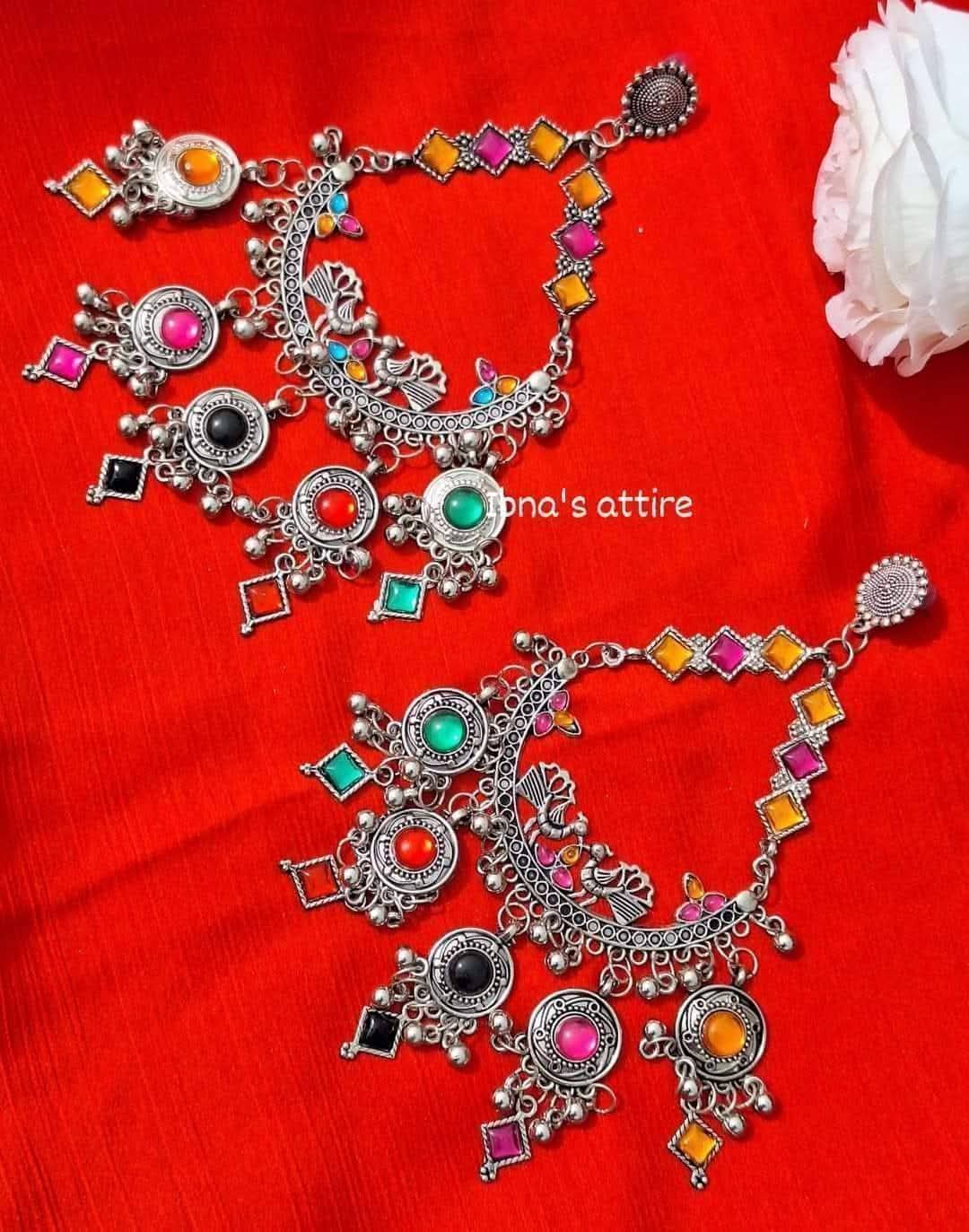 A pair of ornate silver-toned Indian chandbali earrings decorated with colorful rhinestones in pink, orange, yellow, green, black, and red, displayed on a bright red fabric background.