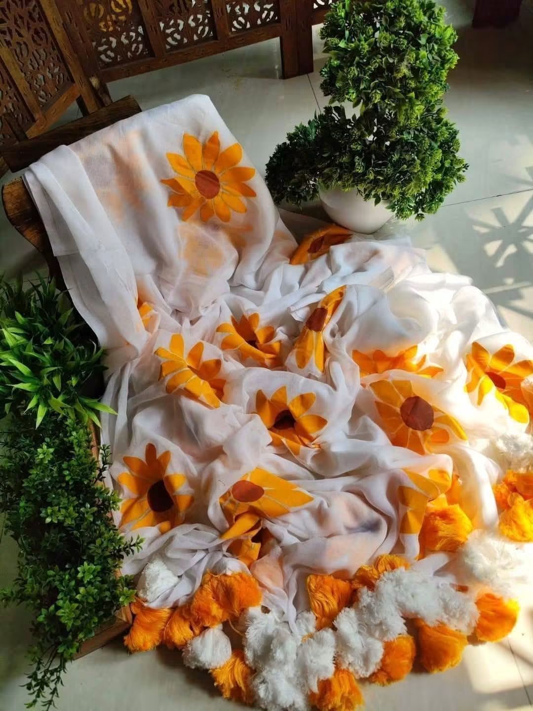 A white hand painted mulmul jandloom saree draped over a chair, decorated with large orange and yellow sunflower motifs and fluffy orange pom-pom trim, styled indoors with potted green plants.