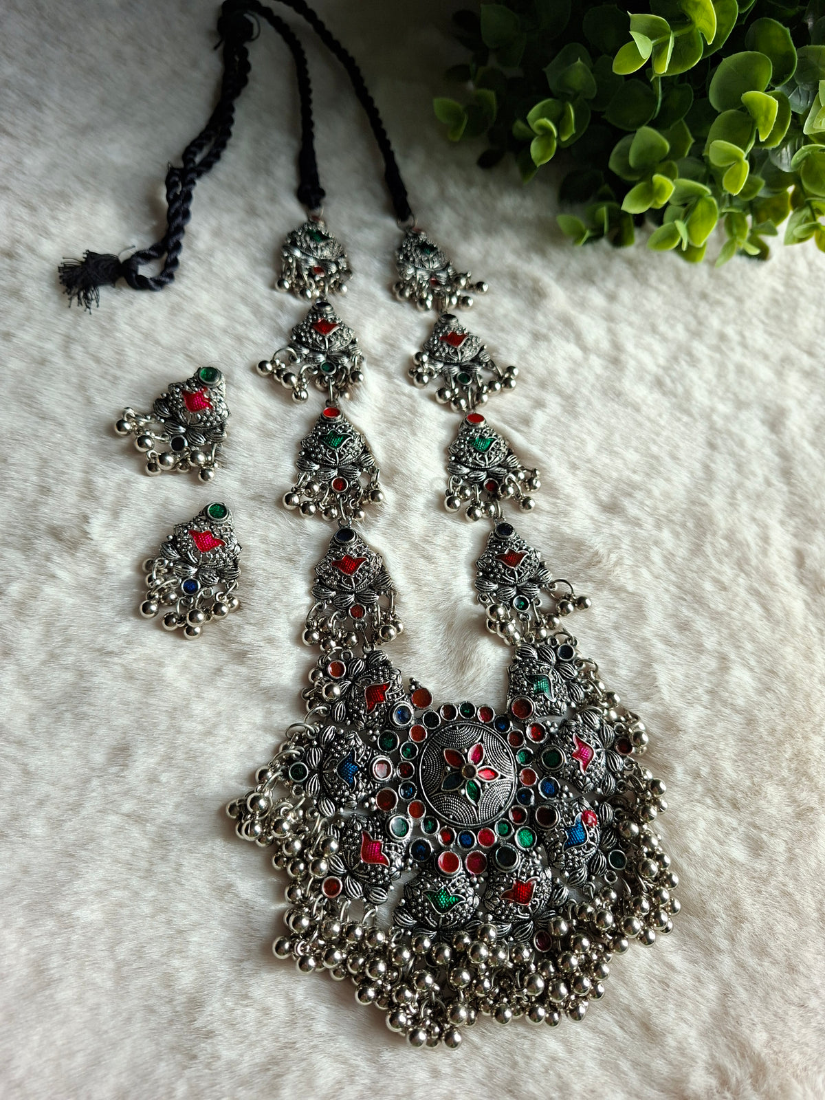 Featuring a German Silver Oxidized Long Necklace Set. Fine traditional motifs pressed into the silver alloy for an authentic antique look.Accents of rich ruby-red, emerald-green, and sapphire-blue stones scattered across the pendant and side links.Abundant tiny silver bells ringing softly and adding heavy structural movement along the lower edges.