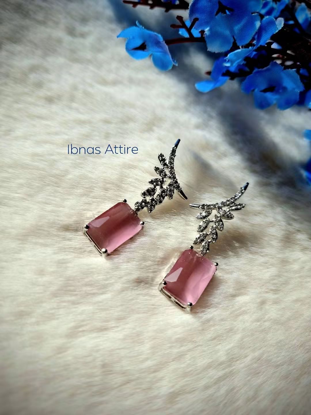 Silver-tone drop earrings with diamond-encrusted leaf posts and rectangular pink stone drops, displayed on a soft white fabric background with blue flower decor.