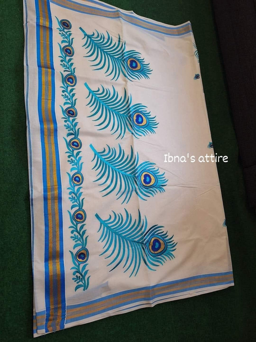 Folded off-white pure cotton Kerala Kasavu saree showcasing woven gold-and-blue border stripes alongside detailed blue embroidered peacock feathers.Lightweight and breathable fabric ensuring all-day comfort during festive gatherings and weddings.Traditional gold and blue woven stripes running along the selvage.