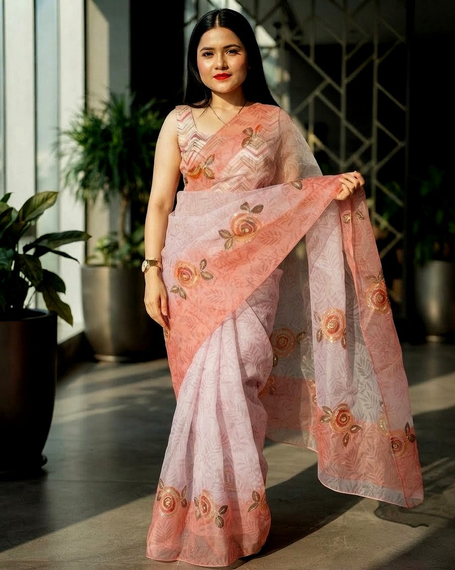 A woman modeling a peach and light pink leno cotton saree with floral patterns, standing in a modern indoor setting with potted plants.
