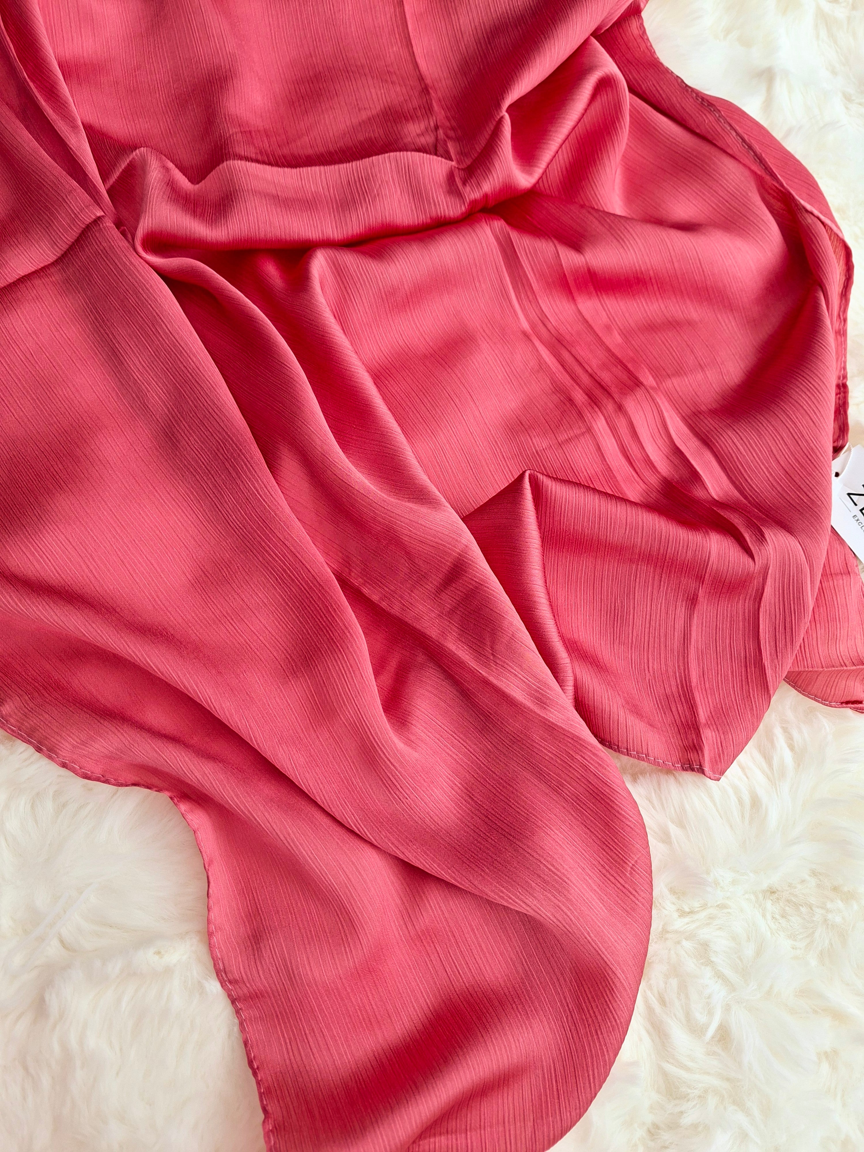 A draped coral pink satin silk hijab, laid out on a white faux fur surface.