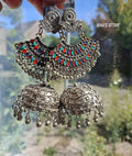 A pair of ornate silver-toned Indian jhumka earrings with intricate filigree work, turquoise and red bead embellishments, and dangling bell-shaped charms, held up by a hand against a blurred outdoor background.