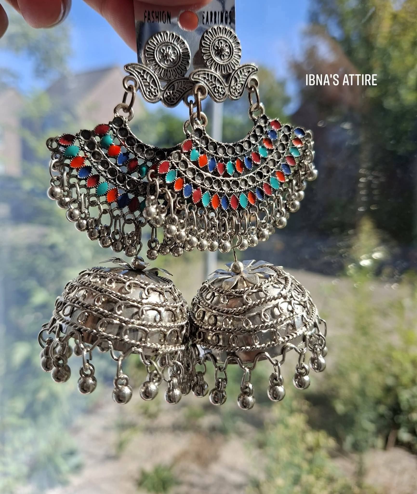 A pair of ornate silver-toned Indian jhumka earrings with intricate filigree work, turquoise and red bead embellishments, and dangling bell-shaped charms, held up by a hand against a blurred outdoor background.