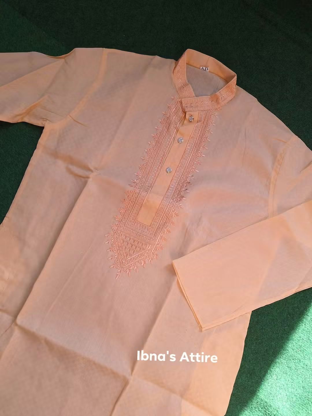 A peach-colored long-sleeve kurta with intricate embroidery on the chest and collar, laid flat on a dark green textured background. The brand name 'Ibna's Attire' is printed in white text near the bottom.