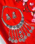 Silver-toned traditional Indian jewelry set featuring a necklace and matching earrings with blue, red, and white enamel details, displayed on a bright red fabric background with pink flowers.