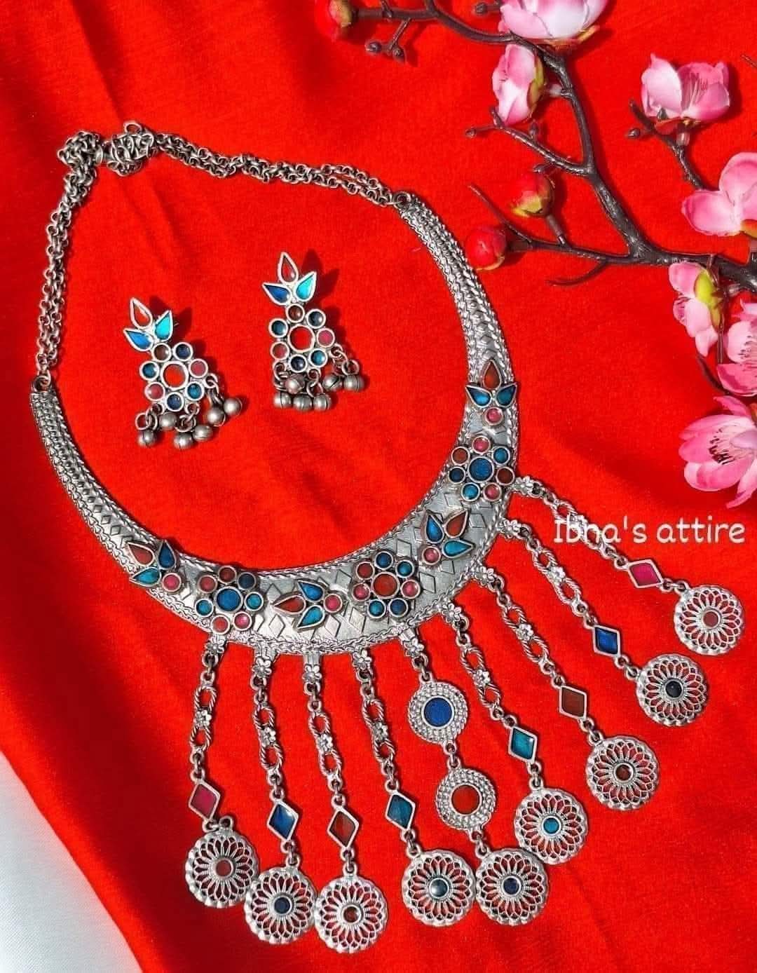 Silver-toned traditional Indian jewelry set featuring a necklace and matching earrings with blue, red, and white enamel details, displayed on a bright red fabric background with pink flowers.