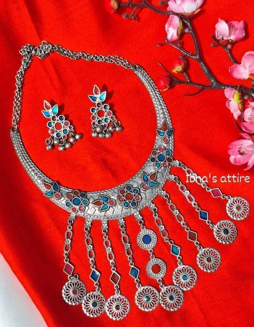 Silver-toned traditional Indian jewelry set featuring a necklace and matching earrings with blue, red, and white enamel details, displayed on a bright red fabric background with pink flowers.