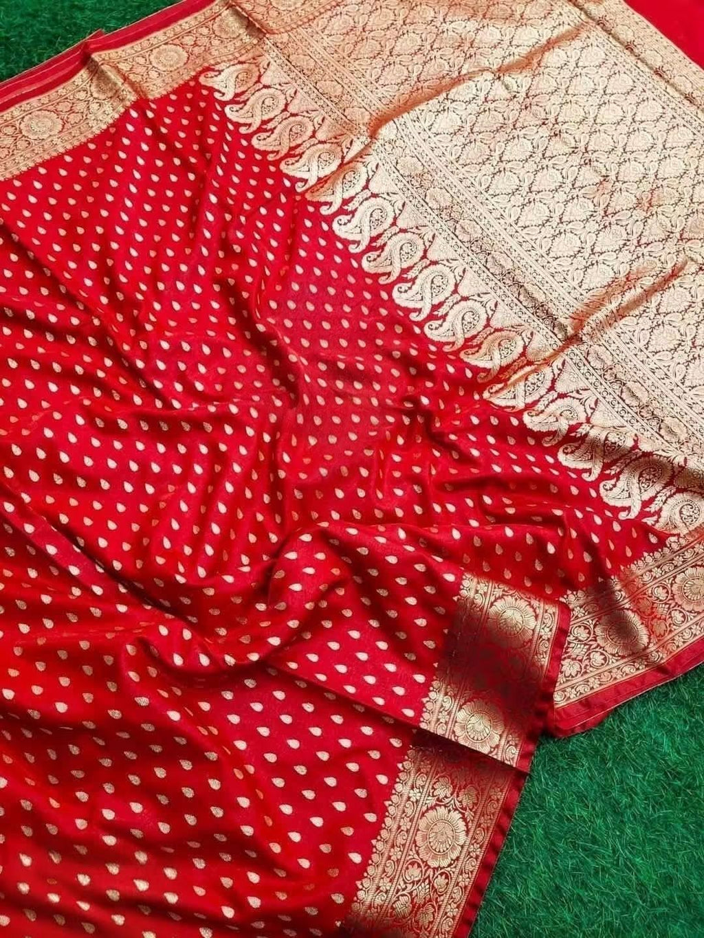 Traditional Banarasi Katan Silk Red saree-Intricate metallic gold jacquard weaving featuring traditional motifs across the body and heavy border framing.Vibrant red dual-tone satin-silk fabric, scattered gold zari bootis, rich gold woven pallu/border resting on green surface.