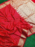 Traditional Banarasi Katan Silk Red saree-Intricate metallic gold jacquard weaving featuring traditional motifs across the body and heavy border framing.Vibrant red dual-tone satin-silk fabric, scattered gold zari bootis, rich gold woven pallu/border resting on green surface.
