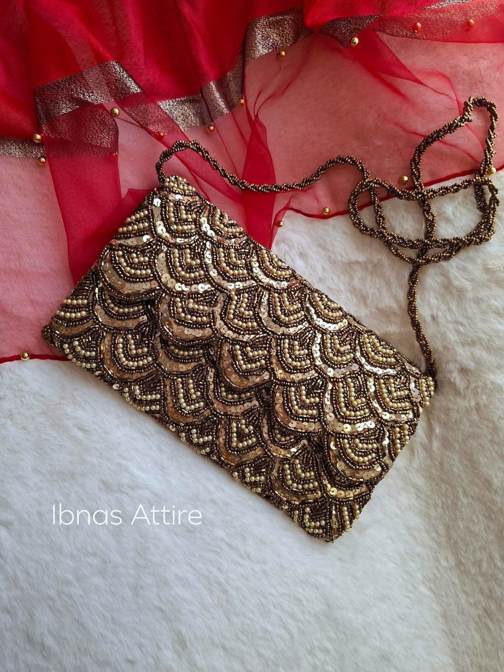 A rectangular clutch bag with an intricate gold and brown scalloped beaded pattern, resting on a white faux fur surface with red sheer fabric in the background. The brand name 'Ibnas Attire' is visible as a watermark in the bottom left corner.