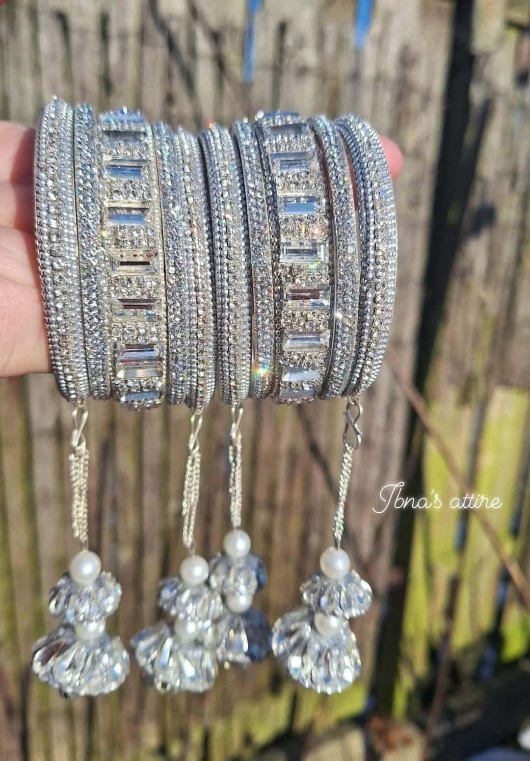 A hand holds a stack of ornate silver-toned bangles decorated with clear crystals and rectangular stone accents, with three dangling crystal clusters and pearl-like beads hanging below. The background is a blurred outdoor wooden fence.