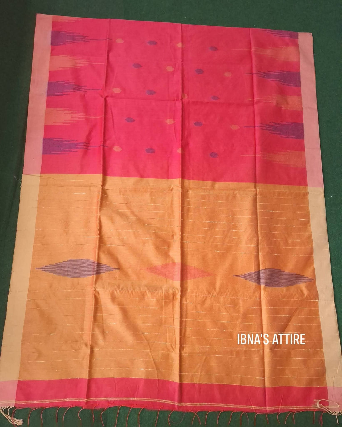 Folded dual-tone saree displayed flat on a green surface, showing upper coral pink half with purple bootis, lower ochre yellow half with horizontal stripes and purple ikat diamonds, edged by a light cream/peach border and bottom fringe tassels.