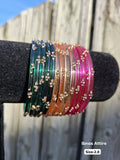 Stack of translucent glass bangles in forest green, champagne amber, and magenta pink with gold sequin accents displayed on a black velvet bar outdoors against a wooden fence.