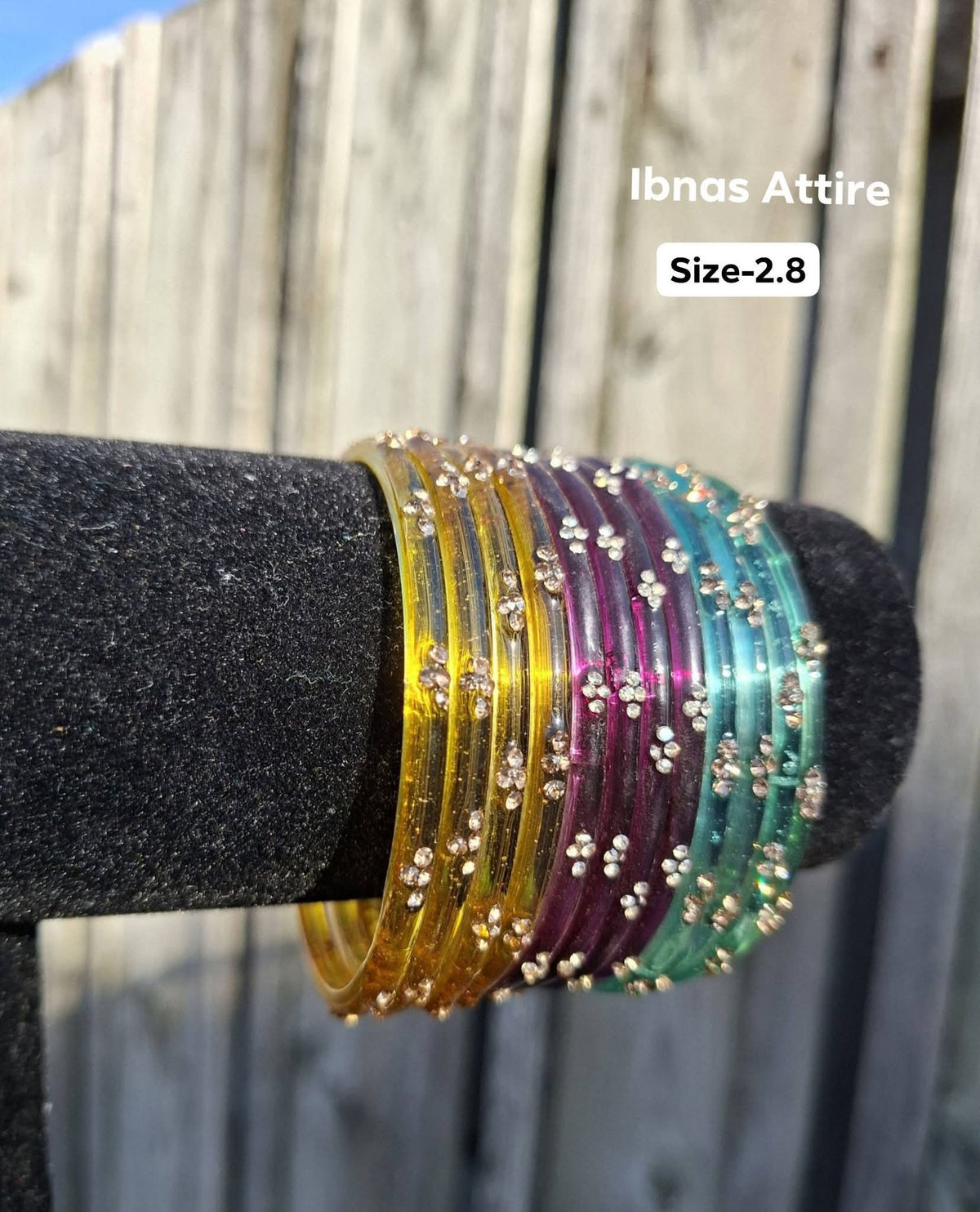 Stack of vibrant multi-colored glass bangles (bright yellow, dark purple, aqua, sea green) with gold sequin accents displayed on a black velvet bar outdoors against a wooden fence.Harmonious blend of sunny yellow, plum purple, and aqua green tones that catch light brilliantly.  