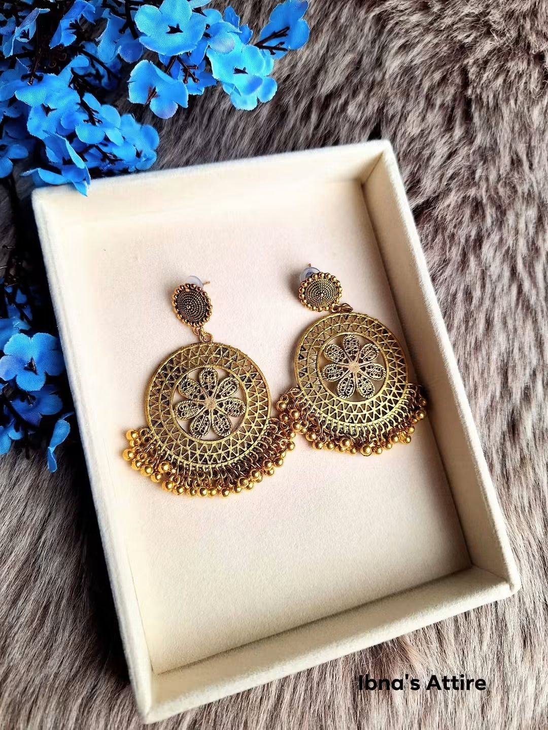 A pair of ornate antique gold drop earrings displayed inside a cream-colored jewelry box, set against a textured gray background with blue flowers in the corner.