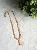 Gold chain necklace with white and ruby-colored zircon clusters, laid out on a white textured background next to green foliage.