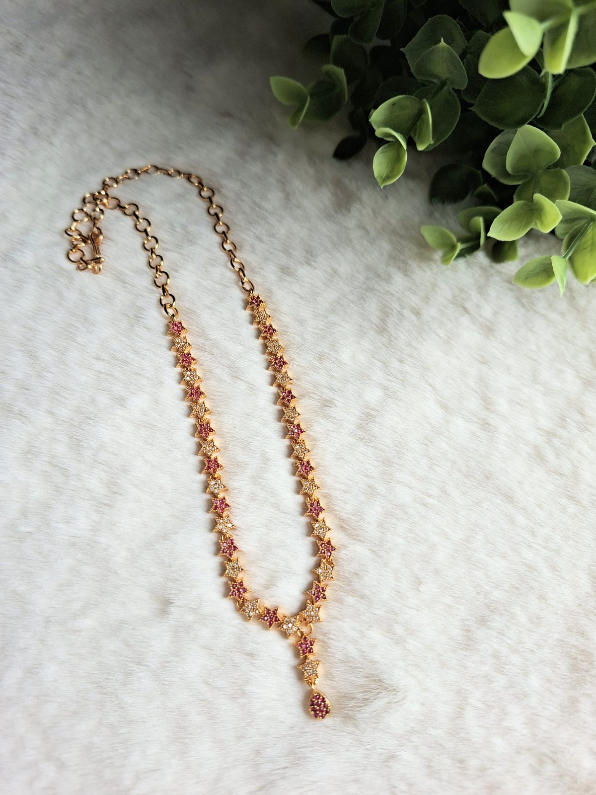 Gold chain necklace with white and ruby-colored zircon clusters, laid out on a white textured background next to green foliage.