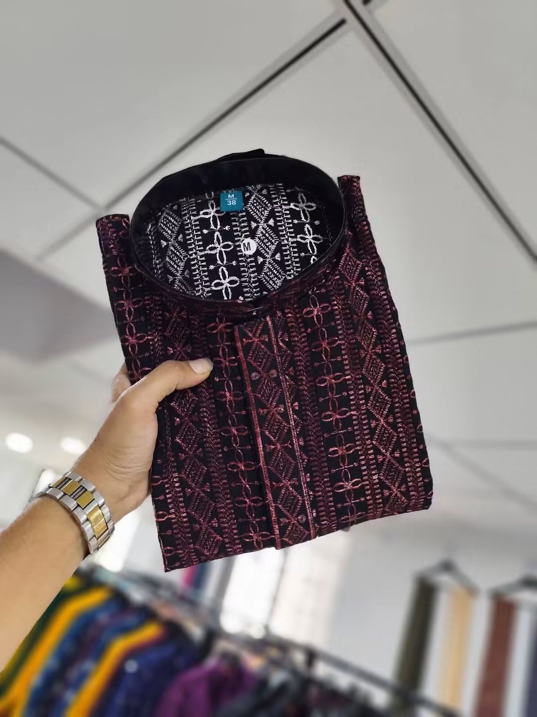 A hand holds up a folded patterned shirt or tunic in a retail store setting. The garment has a dark base with intricate pink and white geometric and floral embroidery.