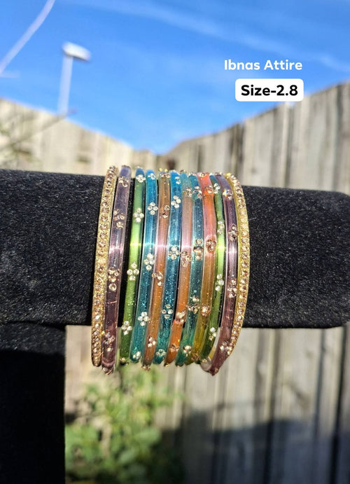 Stack of multi-colored translucent glass bangles (purple, green, cyan, orange, teal, pink) with gold sequin clusters and outer gold stone-encrusted bangles displayed on a black velvet bar outdoors against a wooden fence.