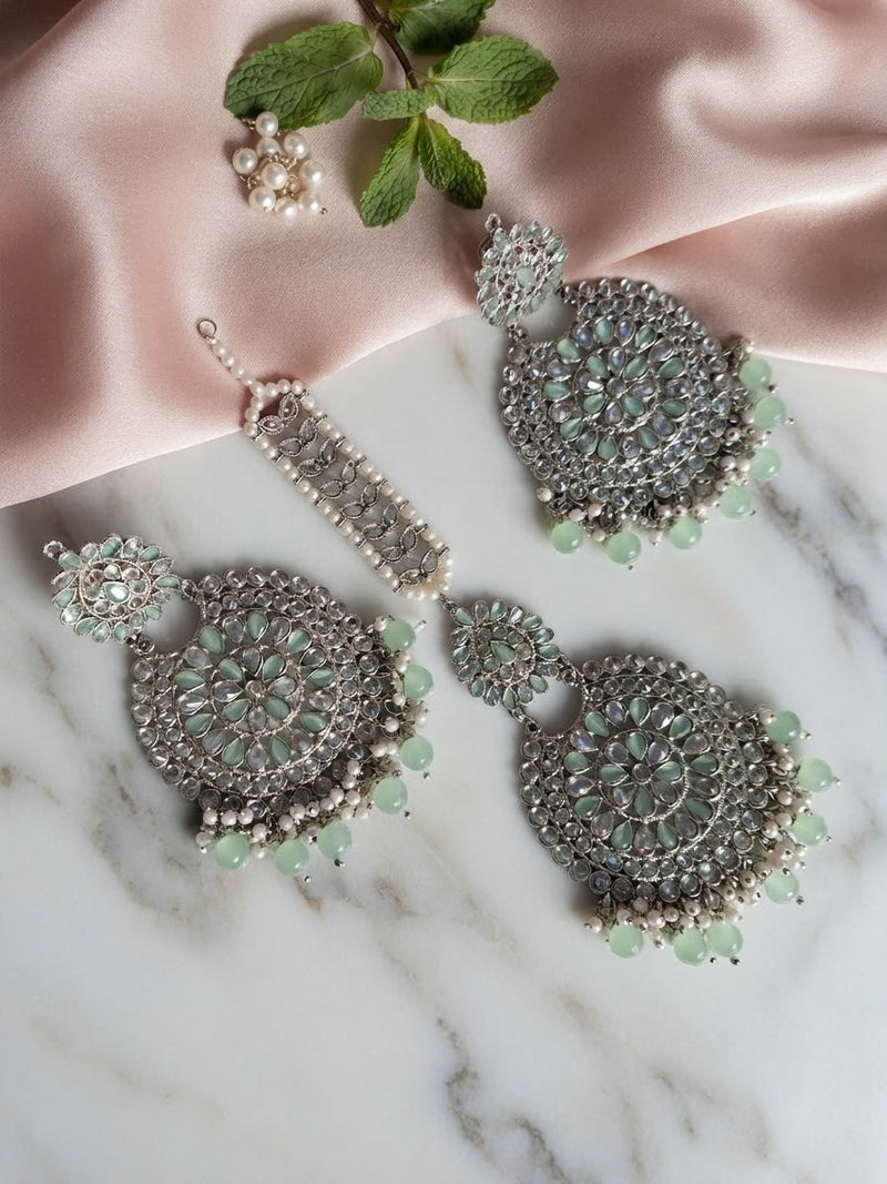 Silver-tone statement earrings and maang tikka featuring reverse AD stones and mint green bead drops, displayed on a satin pinkish fabric background.