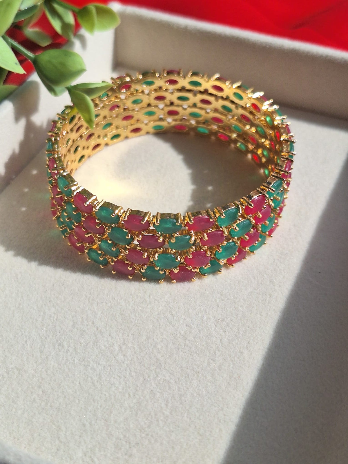 A gold-plated bangle  featuring rows of oval green and pink stones, displayed on a light gray surface with a red background.