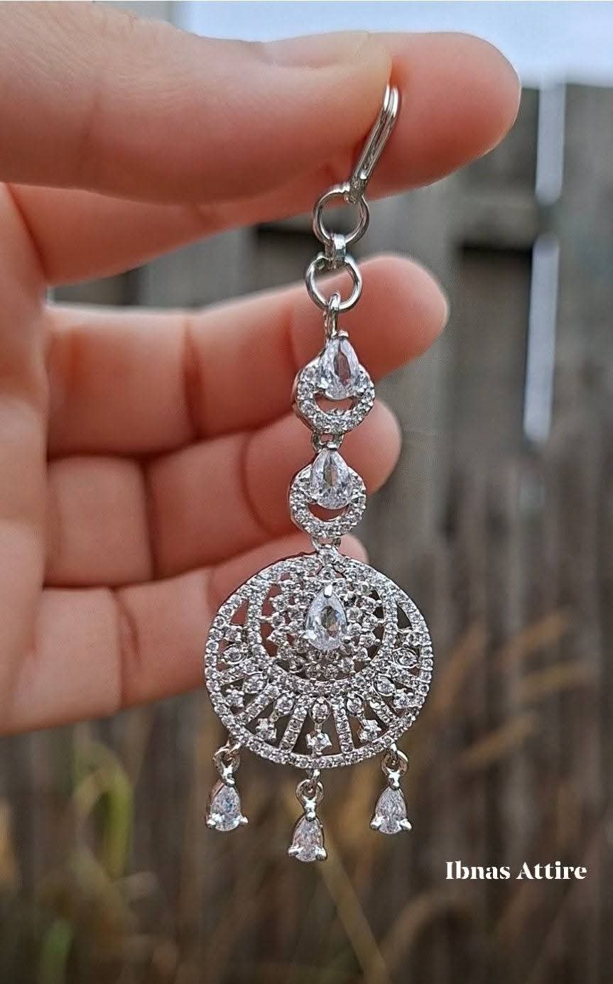 Close-up of a silver American diamond maang tikka featuring clear pave-set stones and teardrop crystal drops held against an outdoor background.High-brilliance stones that capture light from every angle.