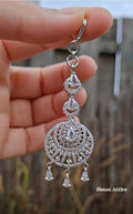 Close-up of a silver American diamond maang tikka featuring clear pave-set stones and teardrop crystal drops held against an outdoor background.High-brilliance stones that capture light from every angle.