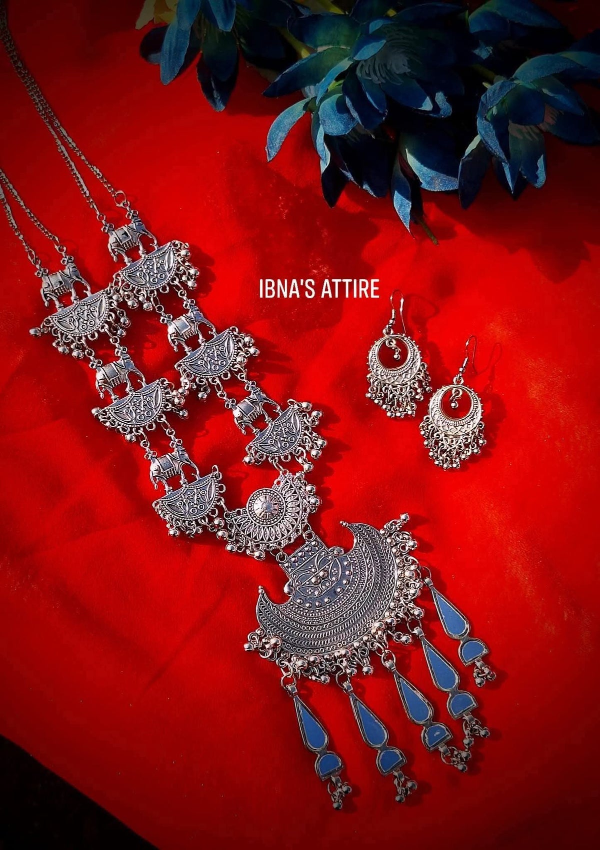 Long oxidized silver chain necklace with elephant links, crescent pendant, mirror charms, matching earrings, laid flat on a red fabric background with blue flowers.