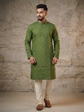 Male model standing against a textured stone wall backdrop wearing an olive green knee-length printed silk kurta with mandarin collar, paired with off-white churidar trousers and brown leather shoes. 