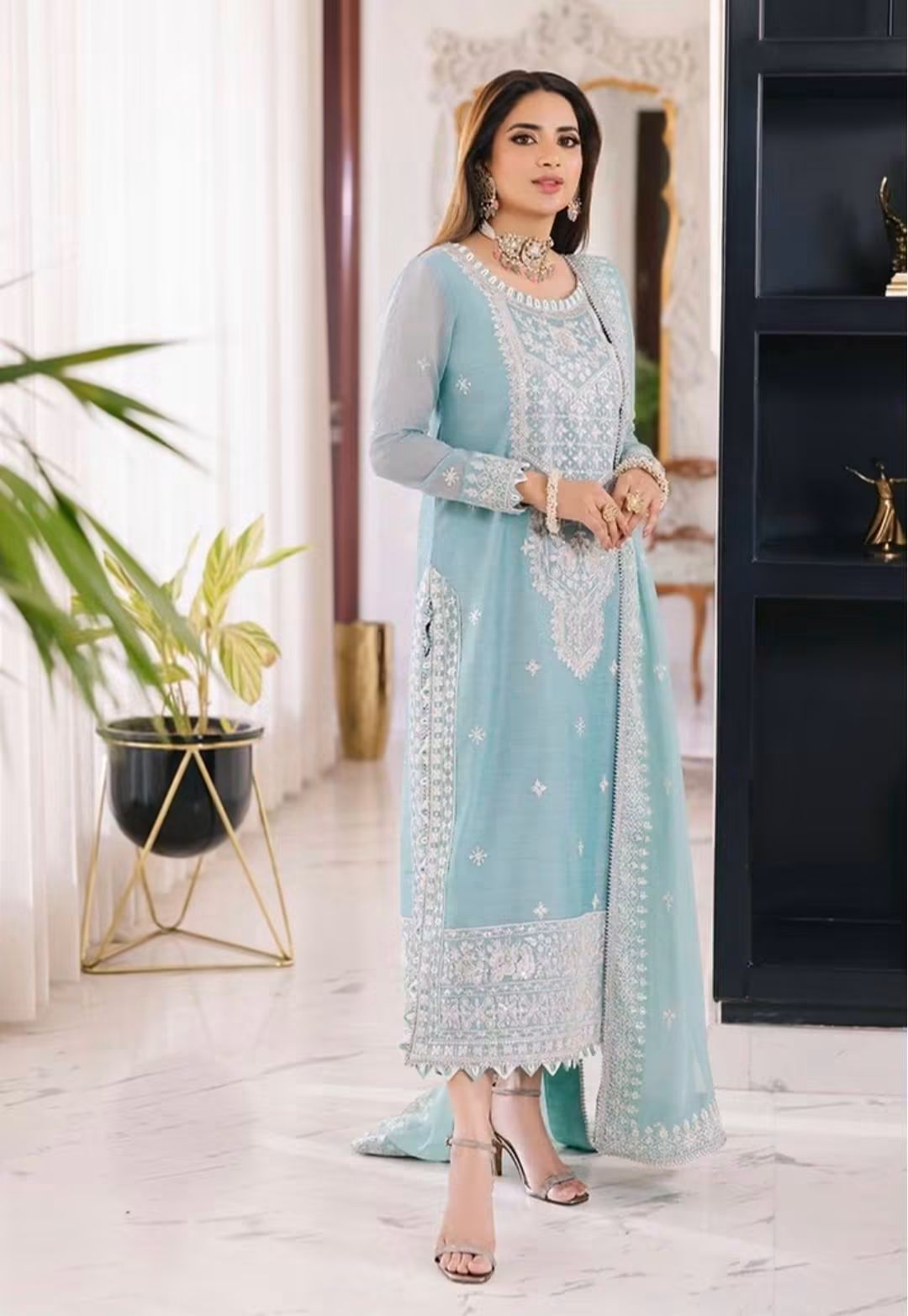A woman models a pastle blue three-piece salwar kameez suit with intricate white embroidery, standing in a modern indoor setting with a marble floor, potted plants, and a dark shelving unit.