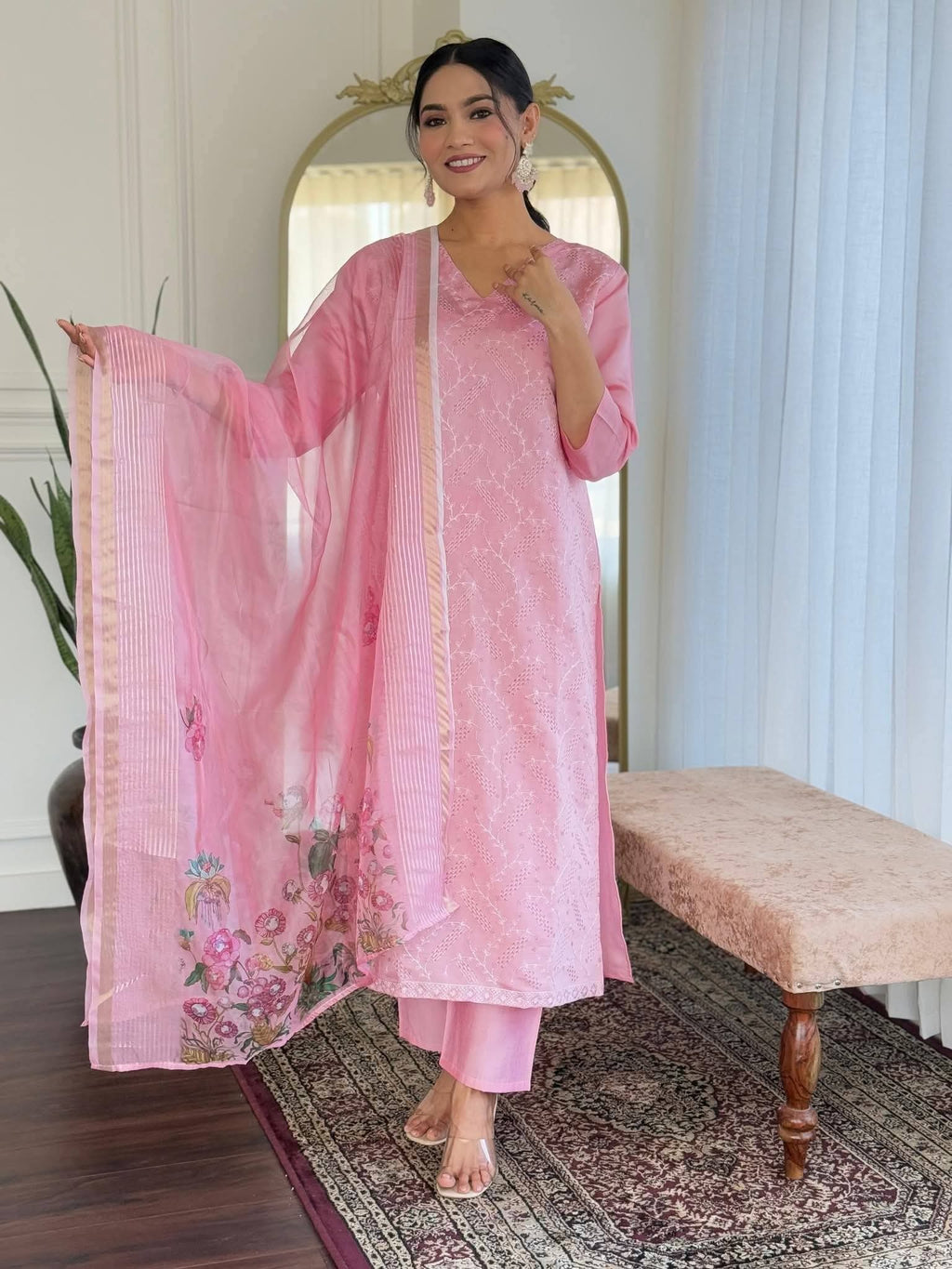 Model wearing a light pink chikan kari sequence-worked Roman silk long kurti and matching trousers, holding a floral digital-printed organza dupatta indoors.High-quality Roman silk offering a structured yet comfortable everyday or party fit.