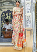  A model wearing an off-white traditional saree with rich multicolored woven borders and pallu, standing against a patterned architectural backdrop.  Intricate all-over woven detailing bringing rich hues to life across the pashmina base.