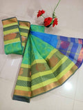 A green and blue patterned karizma silk embossed saree with gold zari borders and motifs, displayed on a light-colored tiled floor next to a small vase of red flowers.