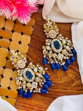 A pair of ornate gold-plated statement earrings with large oval blue crushed stones centers, surrounded by clear stones and dangling blue bead drops, arranged on a wooden surface with decorative fabric and a pink flower.