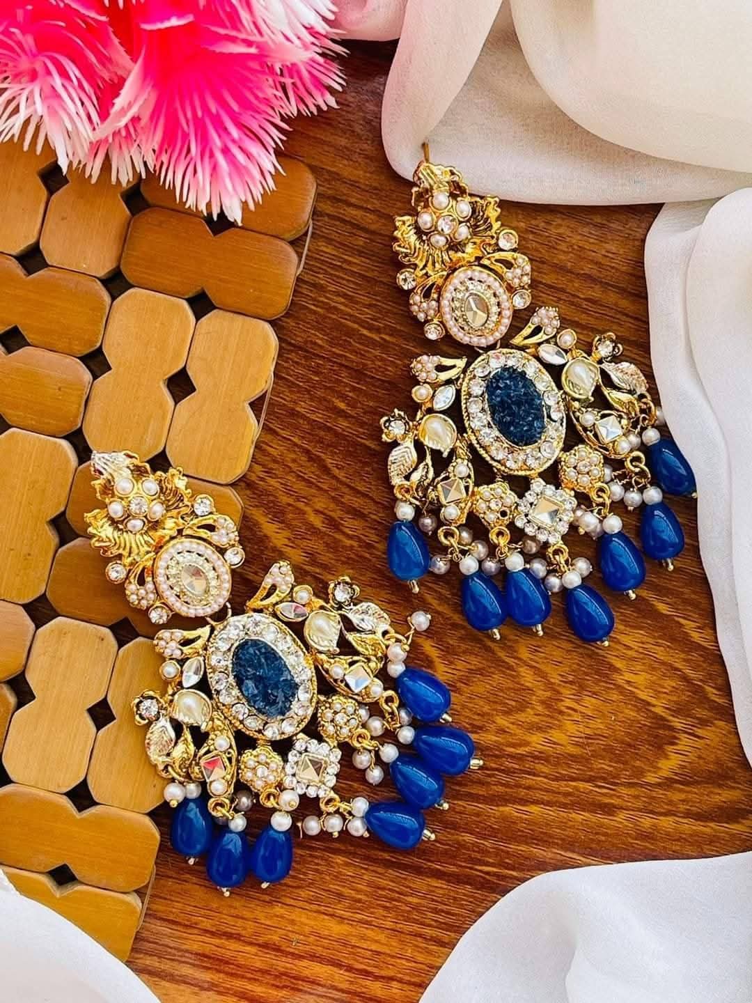 A pair of ornate gold-plated statement earrings with large oval blue crushed stones centers, surrounded by clear stones and dangling blue bead drops, arranged on a wooden surface with decorative fabric and a pink flower.