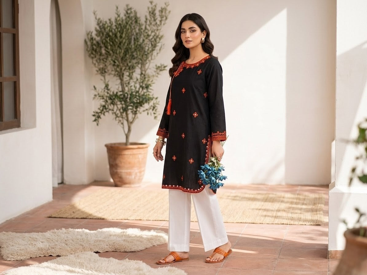 A woman models a black kurta with orange and mirror embroidery, paired with white wide-leg pants, standing in a sunlit courtyard setting.