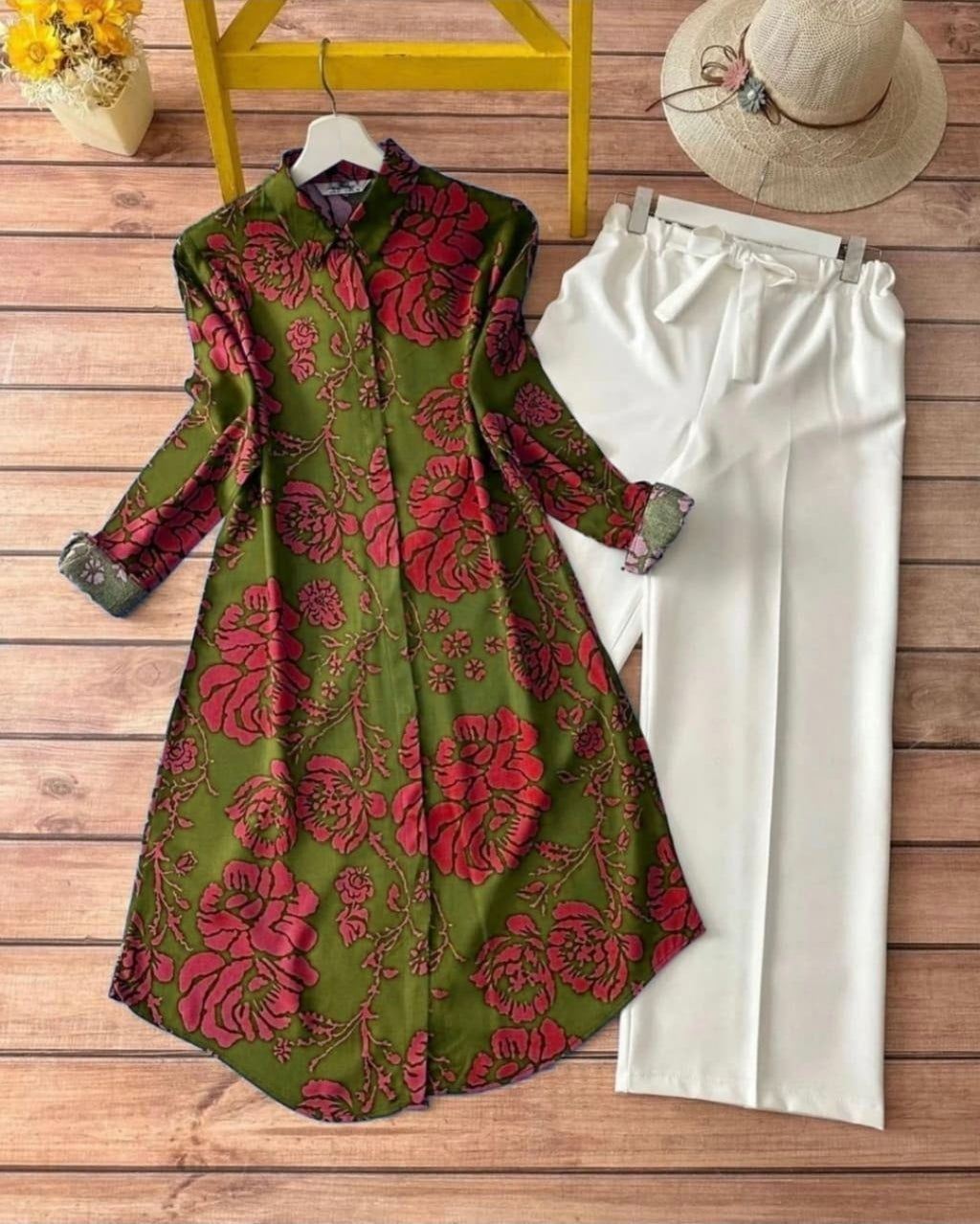 A woman rayon shirt dress in olive green with a bold red floral pattern, styled in a flat lay with white wide-leg trousers, a straw hat, and a small vase of flowers on a wooden floor background.