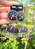Pair of oxidized silver jhumka earrings with multi-color work and ghungroo beads displayed on a backing card against a natural outdoor greenery background. 