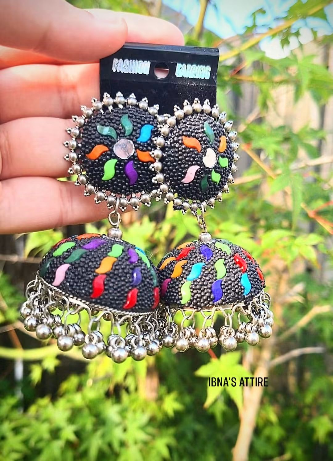 Pair of oxidized silver jhumka earrings with multi-color work and ghungroo beads displayed on a backing card against a natural outdoor greenery background. 