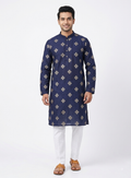 A man modeling a navy blue long-sleeve kurta with white gold geometric patterns, paired with white trousers and brown sandals, against a plain white studio background.