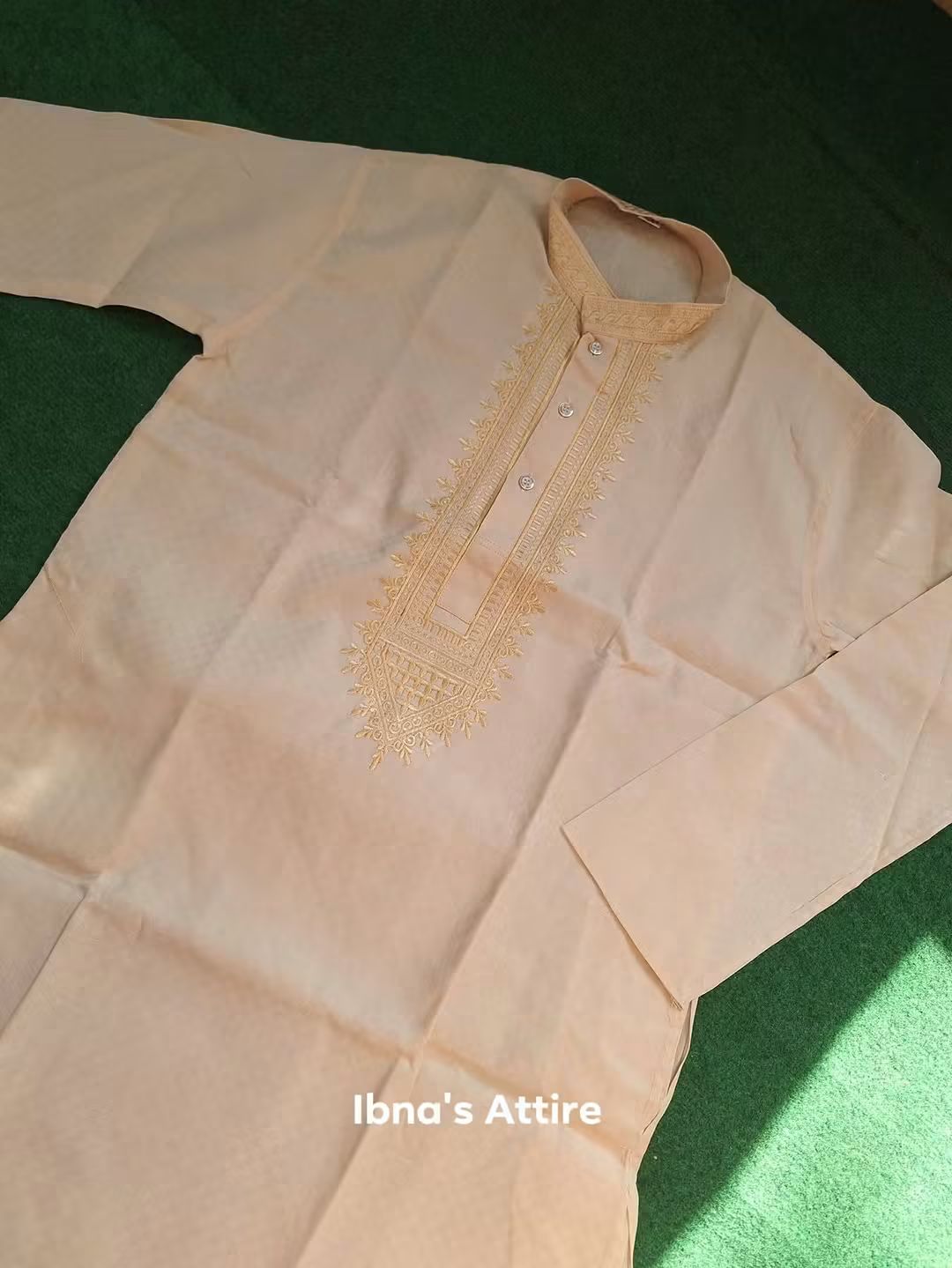A light beige long-sleeve kurta with gold embroidery on the chest, laid flat on a dark green textured background. The brand name 'Ibna's Attire' is printed in white text at the bottom.
